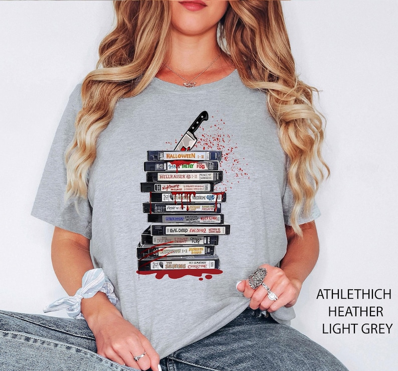 Pode incluir: T-shirt cinzenta clara com um design de cassetes VHS, uma faca e salpicos de sangue. As cassetes t&ecirc;m t&iacute;tulos de filmes como "Halloween" e "Hellraiser". O texto "ATHLETHICH HEATHER LIGHT GREY" est&aacute; impresso no lado direito.
