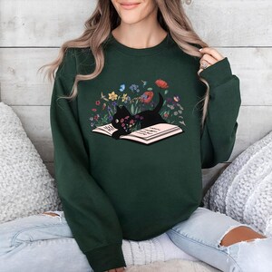 May include: A forest green sweatshirt with a black cat reading a book surrounded by flowers. The cat is lying on the book with its head resting on the pages. The flowers are in shades of pink, purple, yellow, and blue.