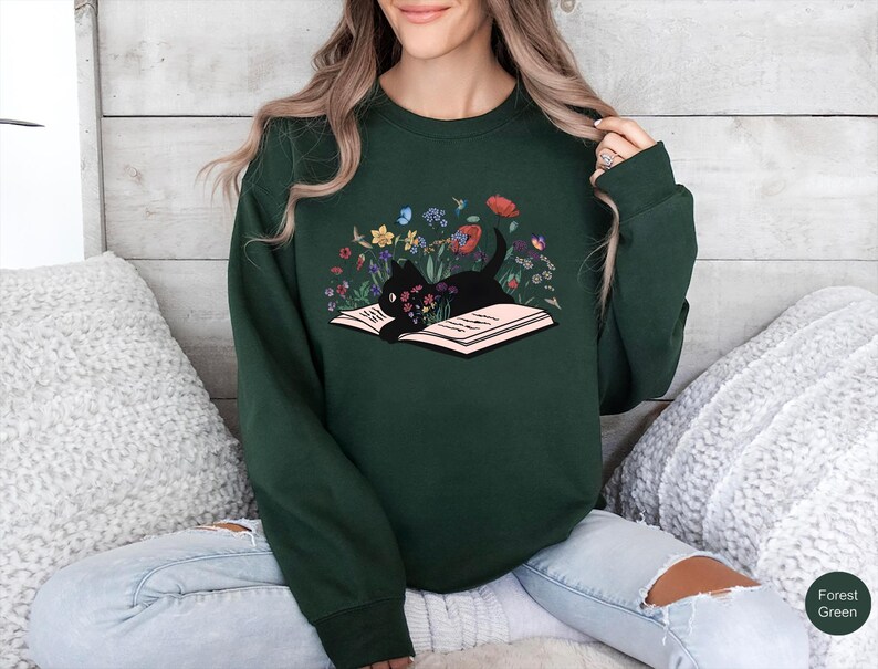 May include: A forest green sweatshirt with a black cat reading a book surrounded by flowers. The cat is lying on the book with its head resting on the pages. The flowers are in shades of pink, purple, yellow, and blue.