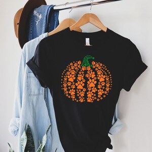 Peut inclure: T-shirt noir avec un motif de citrouille orange compos&eacute; d'empreintes de pattes et d'une tige verte. Le t-shirt est suspendu &agrave; un cintre en bois, avec d'autres v&ecirc;tements en arri&egrave;re-plan.
