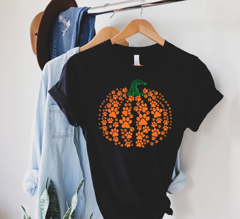 Peut inclure: T-shirt noir avec un motif de citrouille orange compos&eacute; d'empreintes de pattes et d'une tige verte. Le t-shirt est suspendu &agrave; un cintre en bois, avec d'autres v&ecirc;tements en arri&egrave;re-plan.