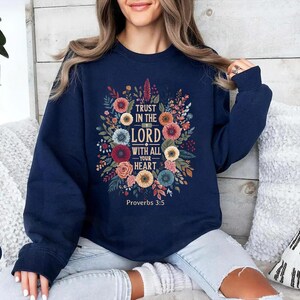 Peut inclure: Sweat-shirt bleu marine avec un motif floral et le texte "Trust in the Lord with all your heart" et le verset biblique Proverbes 3:5. Le motif comprend des fleurs et des feuilles color&eacute;es.