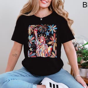 May include: Black t-shirt featuring a colorful illustration of the Statue of Liberty surrounded by flowers and stars. The design is vibrant with red, blue, yellow, and orange hues. The shirt is a classic crew neck style.