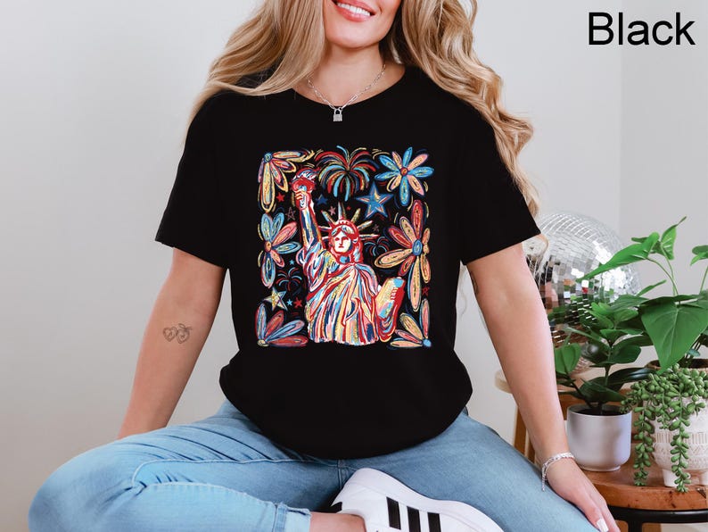 May include: Black t-shirt featuring a colorful illustration of the Statue of Liberty surrounded by flowers and stars. The design is vibrant with red, blue, yellow, and orange hues. The shirt is a classic crew neck style.