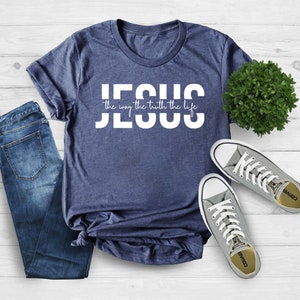 Pu&ograve; includere: Una maglietta blu navy con testo bianco che recita "JESUS the way the truth the life". La maglietta &egrave; stesa su una superficie di legno bianco con un paio di scarpe da ginnastica Converse grigie e una piccola pianta verde.