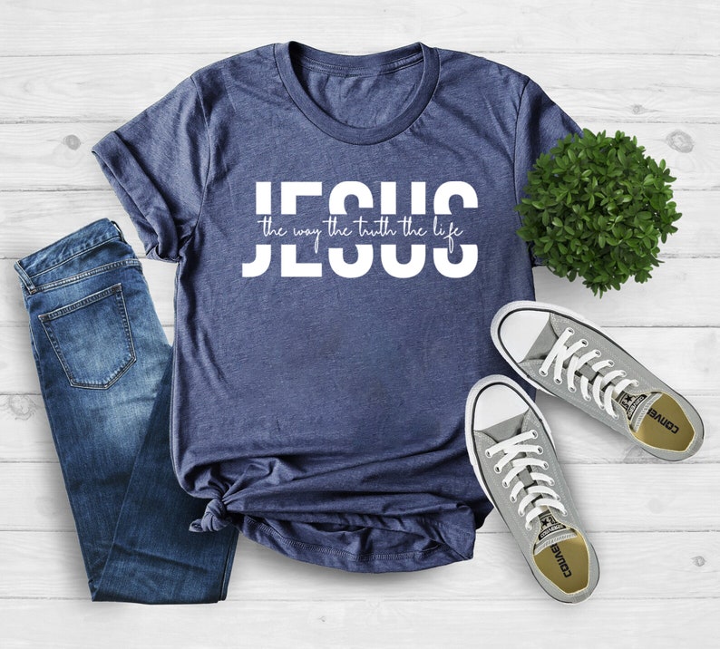 Pu&ograve; includere: Una maglietta blu navy con testo bianco che recita "JESUS the way the truth the life". La maglietta &egrave; stesa su una superficie di legno bianco con un paio di scarpe da ginnastica Converse grigie e una piccola pianta verde.