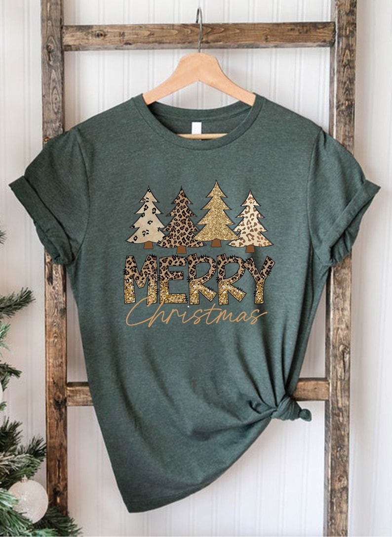May include: A green t-shirt with a graphic of gold glitter and leopard print Christmas trees and the text "Merry Christmas".