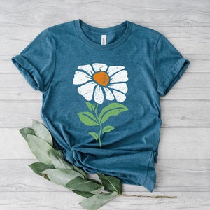 May include: A teal blue t-shirt with a white daisy graphic. The daisy has a yellow center and green leaves.