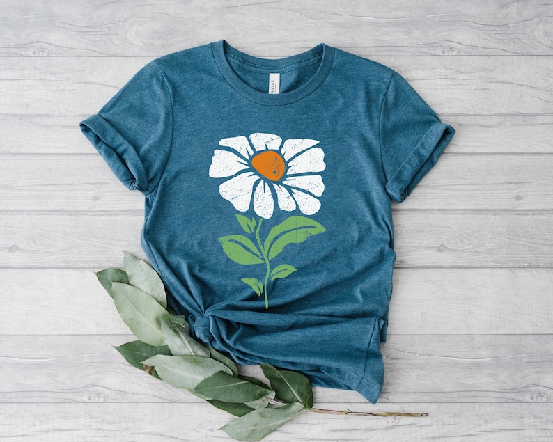 May include: A teal blue t-shirt with a white daisy graphic. The daisy has a yellow center and green leaves.