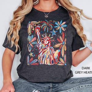 May include: Dark grey heather t-shirt featuring a colorful illustration of the Statue of Liberty, fireworks, and flowers. The design is in a vibrant, abstract style with red, blue, yellow, and orange hues. The text "DARK GREY HEATHER" is printed on the shirt.