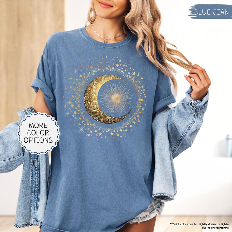 May include: Blue jean-colored t-shirt featuring a gold crescent moon and star design. The moon has an ornate, swirling pattern, and the stars surround the moon in a circular pattern. The shirt has rolled-up sleeves and the text "MORE COLOR OPTIONS" is visible.
