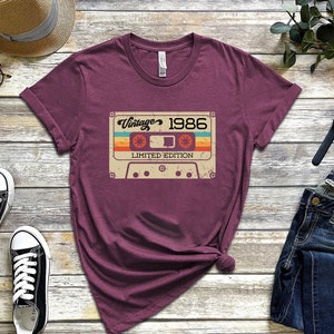 May include: A maroon t-shirt featuring a vintage cassette tape graphic that reads "Vintage 1986 Limited Edition".