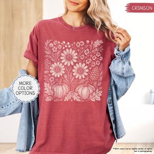 May include: Crimson-colored t-shirt featuring a floral and pumpkin design. The graphic includes large daisies, pumpkins, and various floral elements in a cream color. The shirt has rolled-up sleeves and the text "MORE COLOR OPTIONS" is visible.