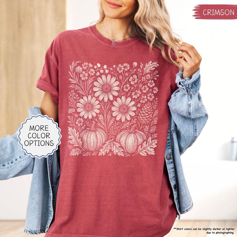 May include: Crimson-colored t-shirt featuring a floral and pumpkin design. The graphic includes large daisies, pumpkins, and various floral elements in a cream color. The shirt has rolled-up sleeves and the text "MORE COLOR OPTIONS" is visible.