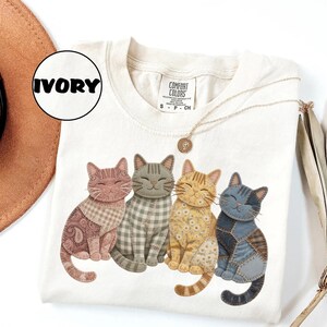 May include: Ivory-colored Comfort Colors t-shirt featuring a graphic of four smiling cats with patchwork patterns. The cats are in shades of brown, gray, yellow, and blue. A gold necklace and a hat are also visible.