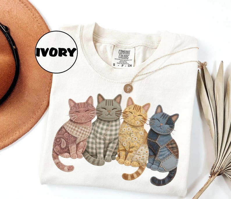 May include: Ivory-colored Comfort Colors t-shirt featuring a graphic of four smiling cats with patchwork patterns. The cats are in shades of brown, gray, yellow, and blue. A gold necklace and a hat are also visible.