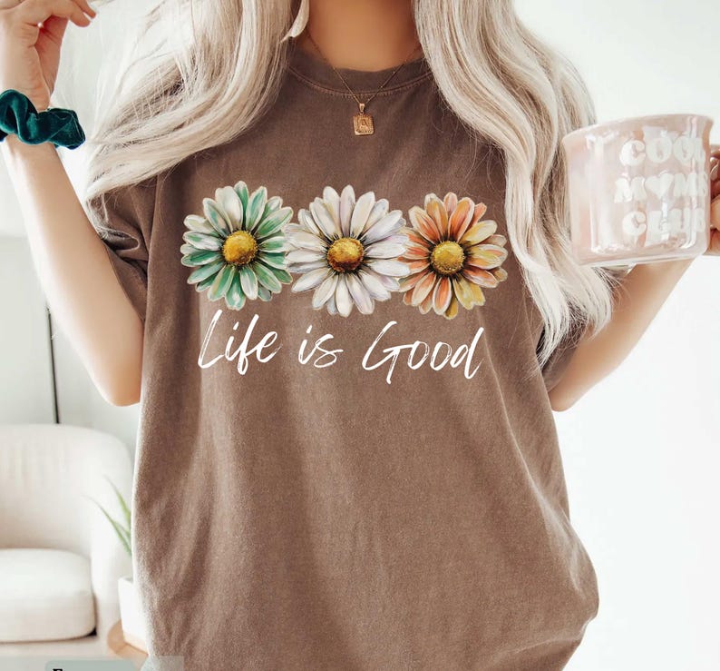 May include: Brown t-shirt featuring three daisy flowers in green, white, and orange with yellow centers. The words "Life is Good" are written in white script below the flowers. The shirt has a vintage, worn look.