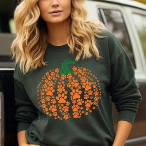 Peut inclure: Sweat-shirt vert fonc&eacute; avec un motif de citrouille compos&eacute; d'empreintes de pattes orange. La tige de la citrouille est faite d'empreintes de pattes vertes. Un design amusant et festif pour les amoureux des animaux, parfait pour l'automne.