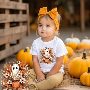 May include: A young child wearing a white t-shirt with a ghost and floral design, paired with mustard-colored pants and a matching headband. The background features pumpkins and a wooden structure, creating a fall theme.