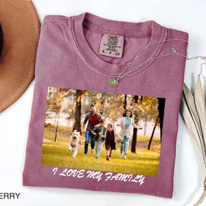 Peut inclure: T-shirt Comfort Colors de couleur baie avec une impression photo de famille. La photo montre une famille courant avec un chien dans un parc. Le texte "I LOVE MY FAMILY" est imprimé en dessous. Un collier argenté est drapé sur le t-shirt.