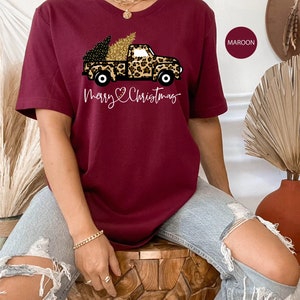 Op de afbeelding: Een maron t-shirt met een afbeelding van een vrachtwagen die een kerstboom vervoert die versierd is met gouden glitter. De vrachtwagen is bedekt met een luipaardprint. De tekst "Merry Christmas" is onder de vrachtwagen gedrukt.