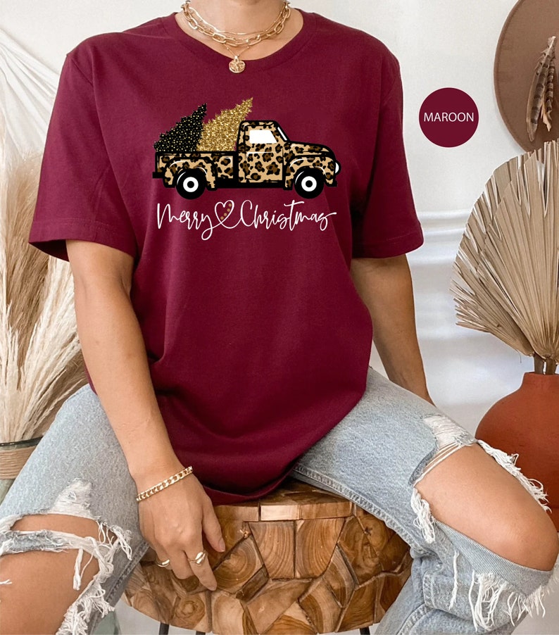 Op de afbeelding: Een maron t-shirt met een afbeelding van een vrachtwagen die een kerstboom vervoert die versierd is met gouden glitter. De vrachtwagen is bedekt met een luipaardprint. De tekst "Merry Christmas" is onder de vrachtwagen gedrukt.