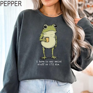 May include: Dark gray sweatshirt featuring a cartoon frog holding a mug. The frog is green and beige, with large eyes. The text "i have to say weird stuff or i'll die" is printed below the frog. The word "PEPPER" is printed at the top left.