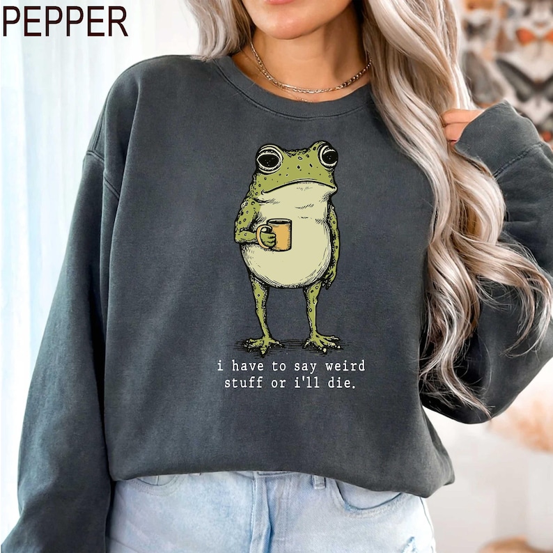 May include: Dark gray sweatshirt featuring a cartoon frog holding a mug. The frog is green and beige, with large eyes. The text "i have to say weird stuff or i'll die" is printed below the frog. The word "PEPPER" is printed at the top left.