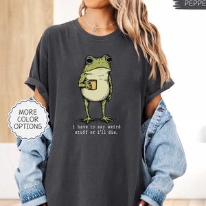 May include: Dark gray t-shirt featuring a cartoon frog holding a mug. The frog is green and white with black outlines. The text on the shirt reads, "I have to say weird stuff or I'll die."