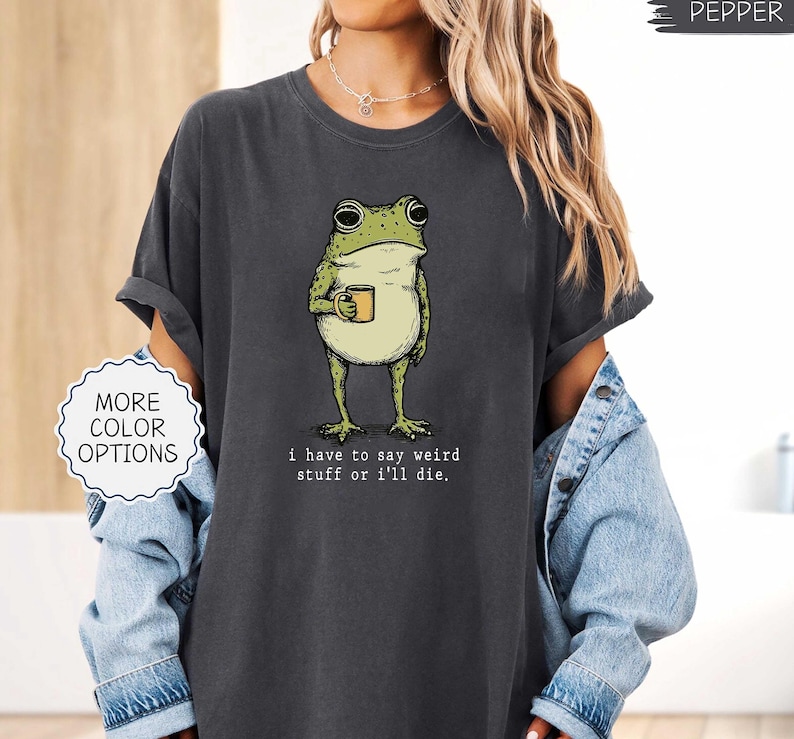 May include: Dark gray t-shirt featuring a cartoon frog holding a mug. The frog is green and white with black outlines. The text on the shirt reads, "I have to say weird stuff or I'll die."