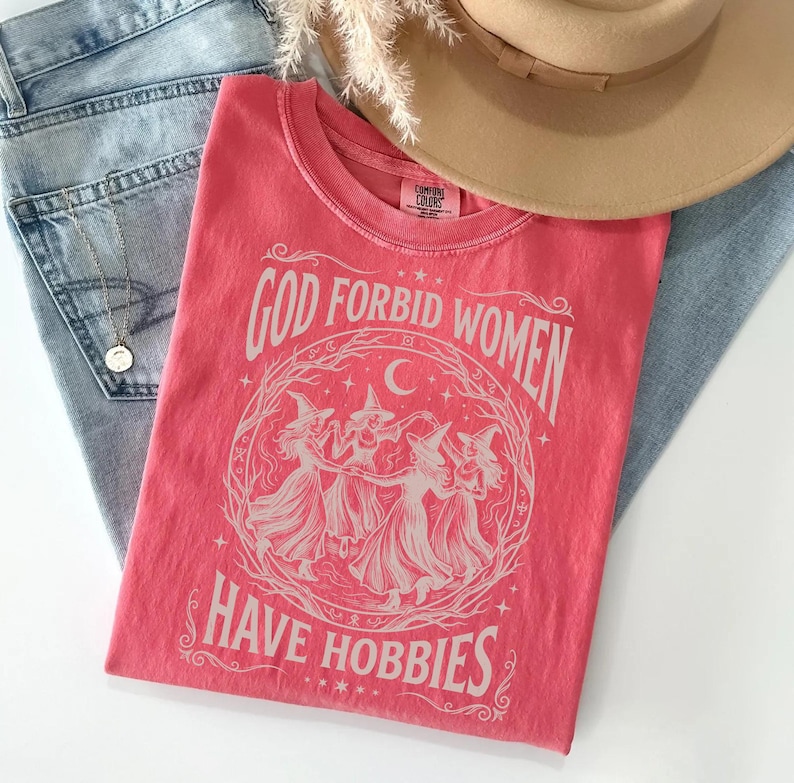 K&ouml;nnte beinhalten: Ein korallenfarbenes T-Shirt mit dem Text "GOD FORBID WOMEN HAVE HOBBIES" und Illustrationen von Hexen. Das T-Shirt liegt gefaltet auf einer blauen Jeans und einem beigen Hut. Marke Comfort Colors.