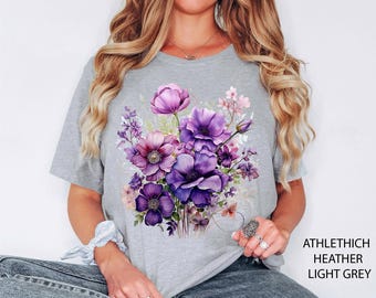 May include: Light grey t-shirt featuring a watercolor floral design in shades of purple and pink. The shirt has short sleeves and a crew neck. The text "ATHLETHICH HEATHER LIGHT GREY" is printed on the right side of the shirt.