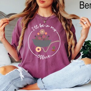 Op de afbeelding: Een paars T-shirt met een witte cirkelvormige afbeelding met een kruiwagen vol bloemen en de tekst "I'll be in my office".