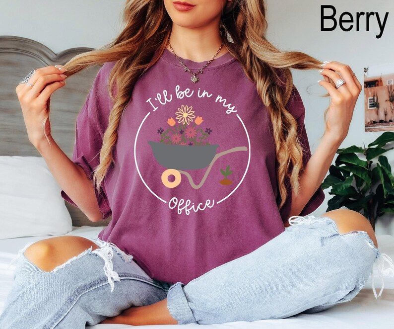 Op de afbeelding: Een paars T-shirt met een witte cirkelvormige afbeelding met een kruiwagen vol bloemen en de tekst "I'll be in my office".