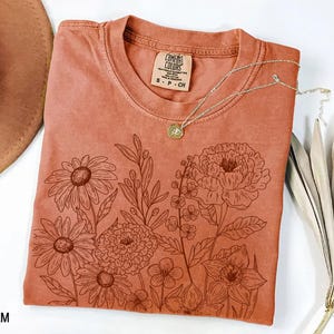 K&ouml;nnte beinhalten: Ein terrakottafarbenes T-Shirt mit floralem Design. Die Grafik enth&auml;lt detaillierte Strichzeichnungen verschiedener Blumen und Bl&auml;tter. Eine silberne Halskette mit Anh&auml;nger liegt auf dem Shirt. Das Shirt hat ein Comfort Colors-Etikett.