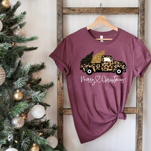 Op de afbeelding: Een maroon t-shirt met een afbeelding van een vrachtwagen die een kerstboom vervoert. De vrachtwagen is bedekt met een luipaardprint met gouden glitteraccenten. De tekst "Merry & Christmas" is onder de vrachtwagen gedrukt.