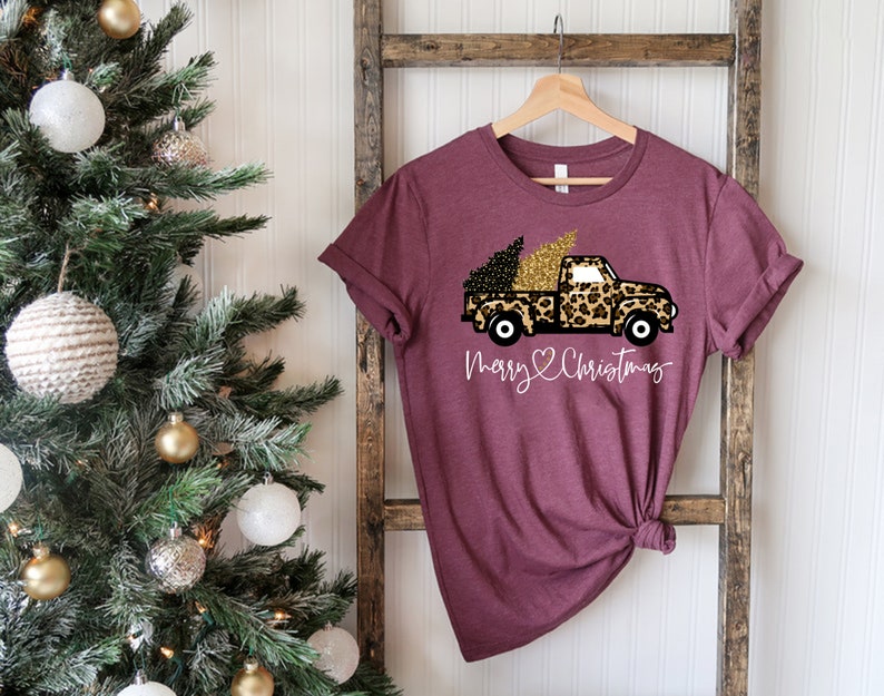 Op de afbeelding: Een maroon t-shirt met een afbeelding van een vrachtwagen die een kerstboom vervoert. De vrachtwagen is bedekt met een luipaardprint met gouden glitteraccenten. De tekst "Merry & Christmas" is onder de vrachtwagen gedrukt.