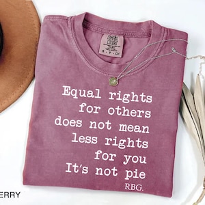May include: Berry-colored t-shirt with the text "Equal rights for others does not mean less rights for you. It's not pie. RBG." A silver necklace with a pendant is draped on the shirt. The shirt is folded on a white surface.