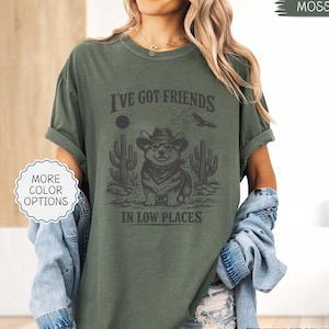 May include: Moss green t-shirt with a graphic featuring a corgi wearing a cowboy hat, cacti, and the text "I've Got Friends In Low Places." A white and blue "More Color Options" button is on the left.