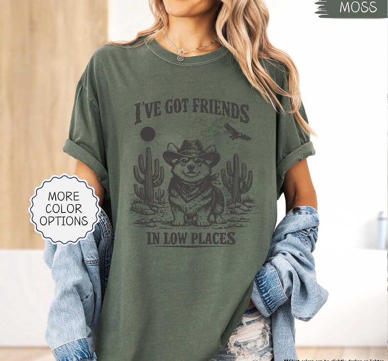 May include: Moss green t-shirt with a graphic featuring a corgi wearing a cowboy hat, cacti, and the text "I've Got Friends In Low Places." A white and blue "More Color Options" button is on the left.