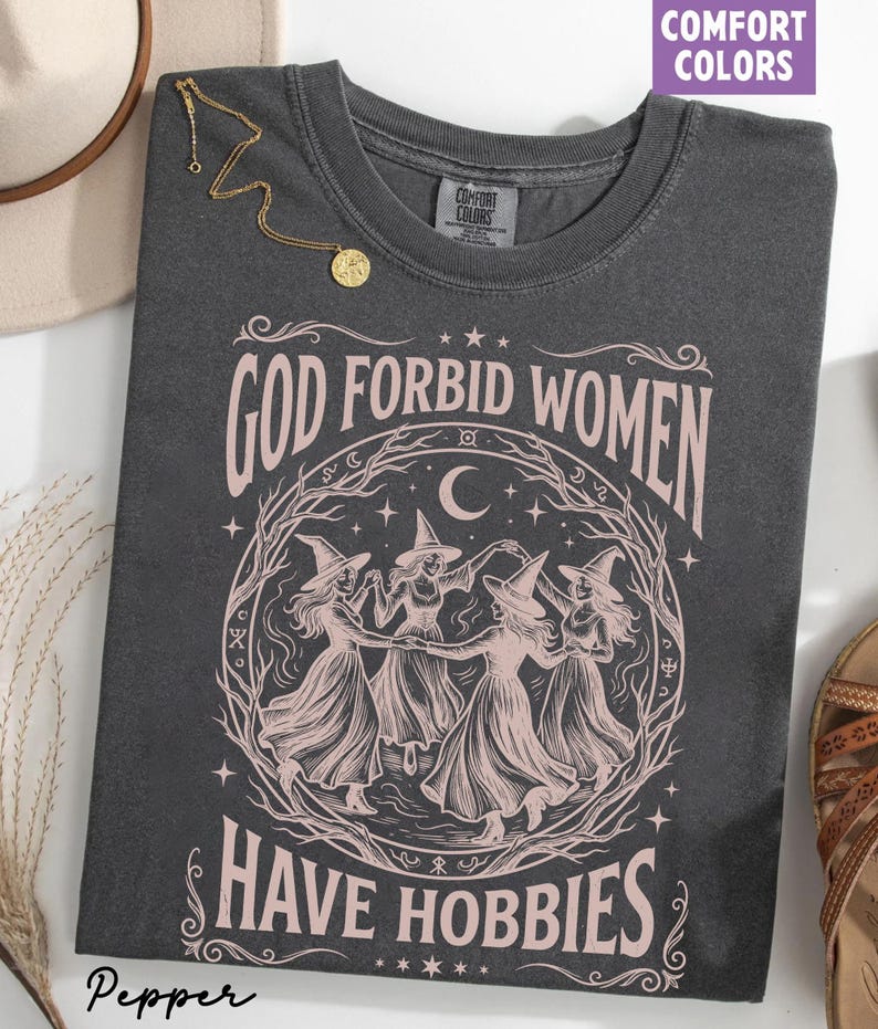 K&ouml;nnte beinhalten: Dunkelgraues T-Shirt mit rosa Aufdruck "GOD FORBID WOMEN HAVE HOBBIES". Die Grafik zeigt vier Hexen, die unter Mond und Sternen im Kreis tanzen. Marke Comfort Colors.
