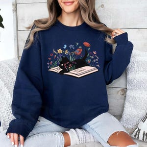 May include: A navy blue sweatshirt with a black cat reading a book surrounded by flowers. The cat is lying on the open book. The flowers are in shades of pink, red, yellow, and blue.