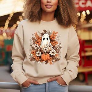 May include: Beige sweatshirt featuring a Halloween-themed graphic. The design includes a white ghost with black eyes, surrounded by orange and white flowers, brown leaves, and a small orange pumpkin. The sweatshirt is a neutral color.