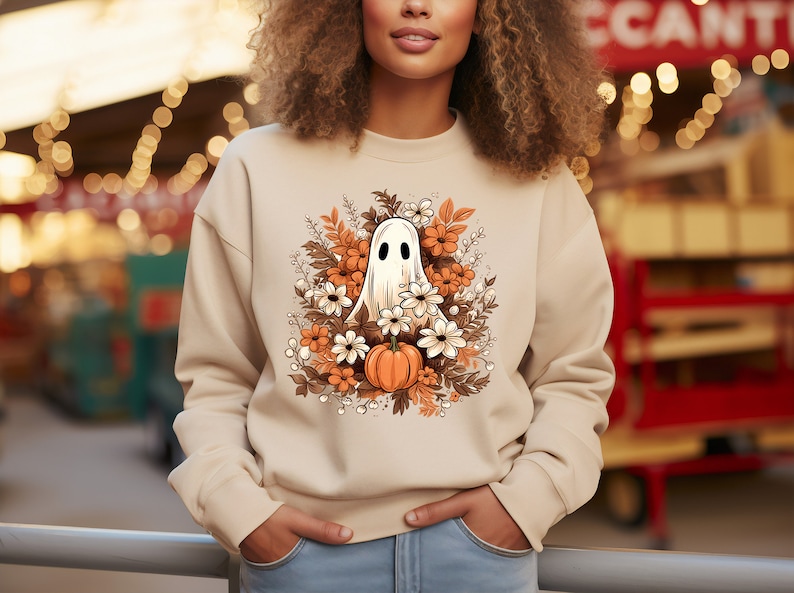 May include: Beige sweatshirt featuring a Halloween-themed graphic. The design includes a white ghost with black eyes, surrounded by orange and white flowers, brown leaves, and a small orange pumpkin. The sweatshirt is a neutral color.