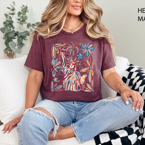May include: Heather maroon t-shirt featuring a colorful illustration of the Statue of Liberty surrounded by flowers, stars, and fireworks. The shirt has a v-neck and short sleeves. The text "HEATHER MAROON" is printed on the upper right corner.
