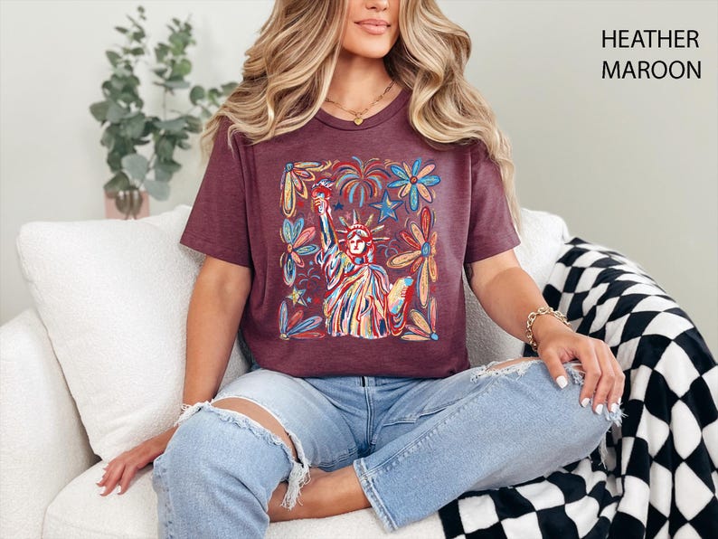 May include: Heather maroon t-shirt featuring a colorful illustration of the Statue of Liberty surrounded by flowers, stars, and fireworks. The shirt has a v-neck and short sleeves. The text "HEATHER MAROON" is printed on the upper right corner.