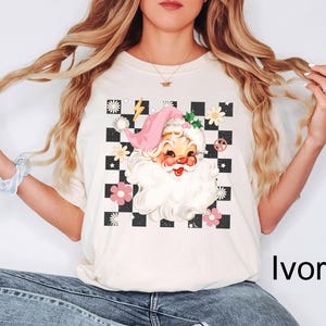 May include: Ivory-colored t-shirt featuring a retro Santa Claus graphic. The design includes a pink hat, a checkered background, flowers, and a peace sign. The text "Ivory" is printed on the bottom right.