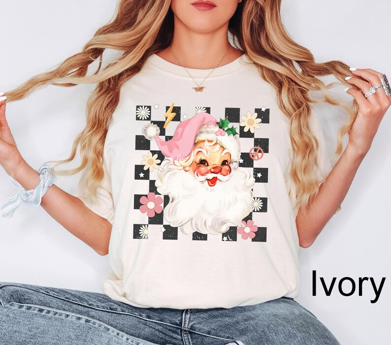 May include: Ivory-colored t-shirt featuring a retro Santa Claus graphic. The design includes a pink hat, a checkered background, flowers, and a peace sign. The text "Ivory" is printed on the bottom right.