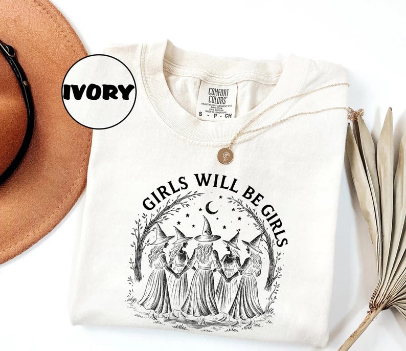 K&ouml;nnte beinhalten: Elfenbeinfarbenes Sweatshirt mit dem Text "GIRLS WILL BE GIRLS" in Schwarz. Die Grafik zeigt f&uuml;nf Figuren in Hexenh&uuml;ten und Kleidern, umgeben von einem Halbmond, Sternen und einem Astmuster. Eine goldene Halskette ist &uuml;ber dem Shirt drapiert.