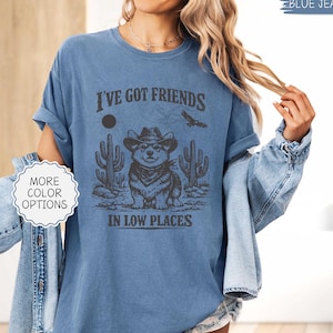 May include: Blue jean-colored t-shirt with a graphic featuring a Corgi wearing a cowboy hat, cacti, and the text "I've Got Friends In Low Places." The shirt has rolled-up sleeves and a relaxed fit.
